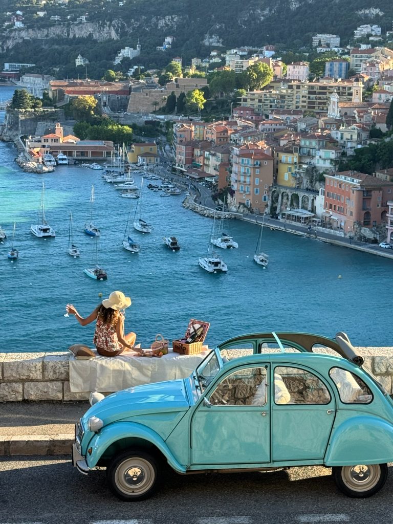 Lady eating a picnic on the edge of Villefranche-sur-mer on the French Riviera by a blue 2CV Citroën car